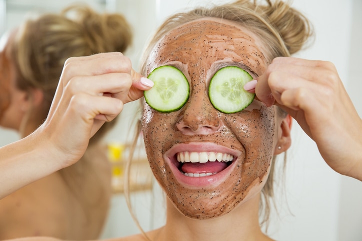 Young beautiful girl applying facial scrub mask on skin. Looking in the mirror in bathroom, Wrapped in a towel, having fun.