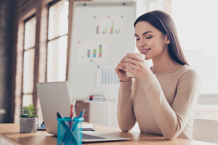 Half-turned portrait of cute calm peaceful feeling comfort coziness pretty tender gentle expert qualified travel agent having rest holding tasty fresh cup of tea in hands sitting at the desktop