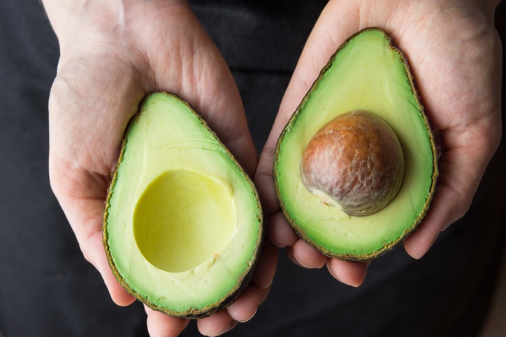 Young Caucasian Woman in Black Apron Holding in Hands Ripe Beautiful Halved Organic Australian Avocado with Pit. Healthy Lifestyle Vegan Vitamin E Oil. Creative Minimalist Food Poster Banner