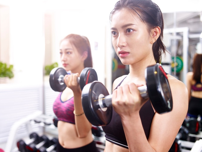 two young asian women working out exercising in gym using dumbbells with serious facial expression.