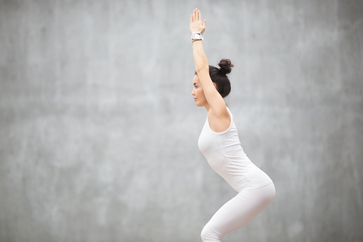 Side view portrait of beautiful young woman wearing white sportswear working out against grey wall, doing yoga or pilates exercise. Standing in Chair, Utkatasana pose. Copy space