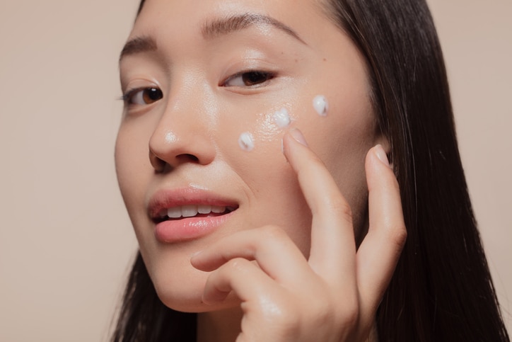 Close up of a young woman applying moisturizer to her face. Asian woman looking happy while following skincare regime.