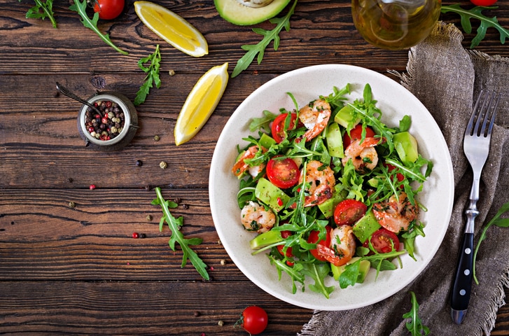 Fresh salad bowl with shrimp, tomato, avocado and arugula on wooden background close up. Healthy food. Clean eating. Top view. Flat lay.