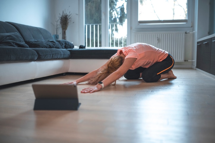 Young woman working out in living room. She is stretching her body in child pose.