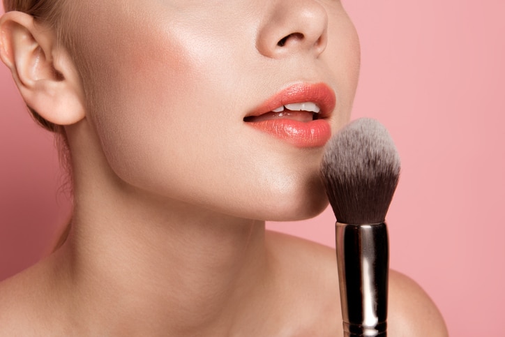 Close up of cared woman face blowing at blush brush. Isolated on rose background