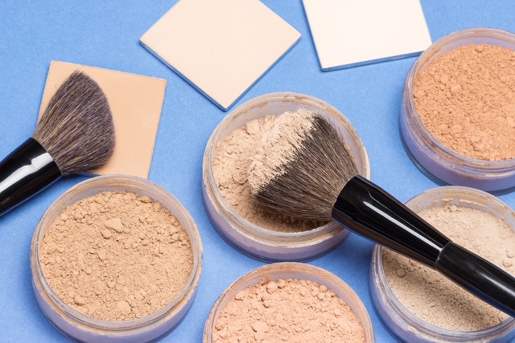 Jars filled with loose cosmetic powder and compact cosmetic powder different shades with makeup brushes on blue background. Focus on central brush