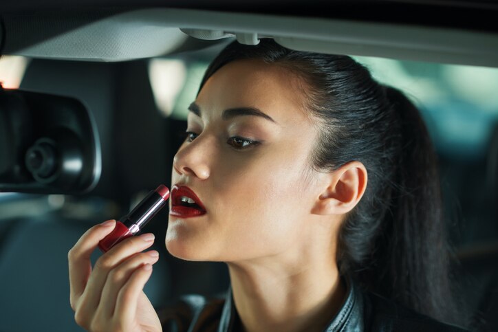 Attractive young woman looking at rearview mirror when applying bright red lipstick