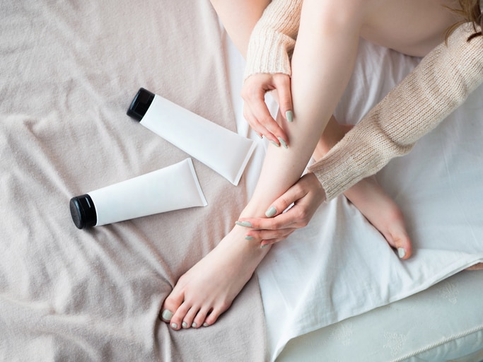 Beautiful young woman applying body lotion on bed sheet. Top view. Blank label for branding mock-up. Woman Skin Care Concept.