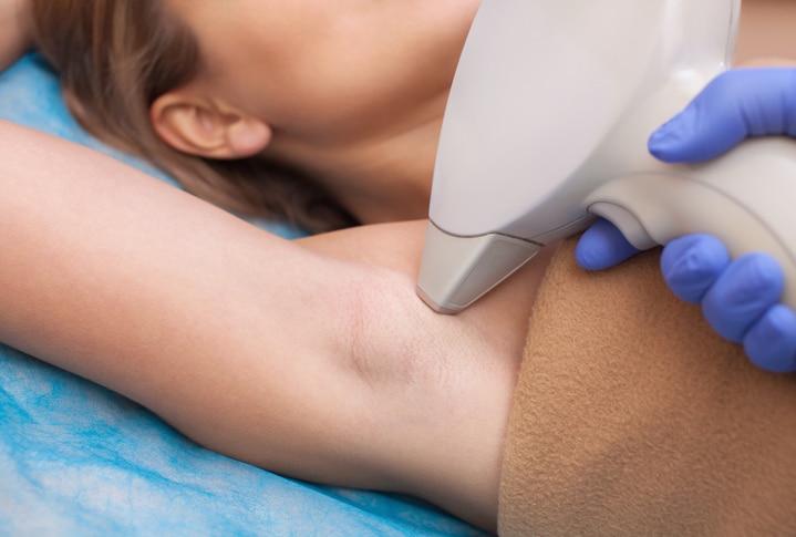 The cosmetologist does the laser hair removal procedure in the armpit zone, to a young woman in a beauty salon.