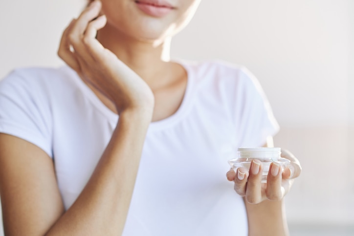 Crop female in white t-shirt taking care of skin putting cream on face on light blur background