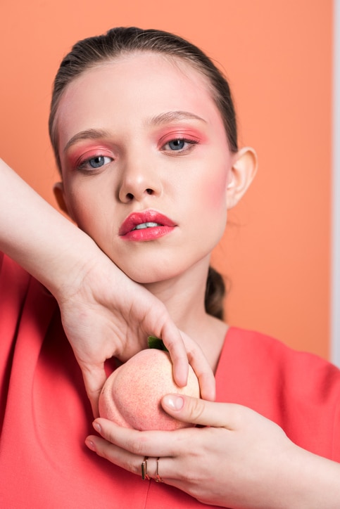 beautiful fashionable girl holding peach, looking at camera and posing with living coral on background