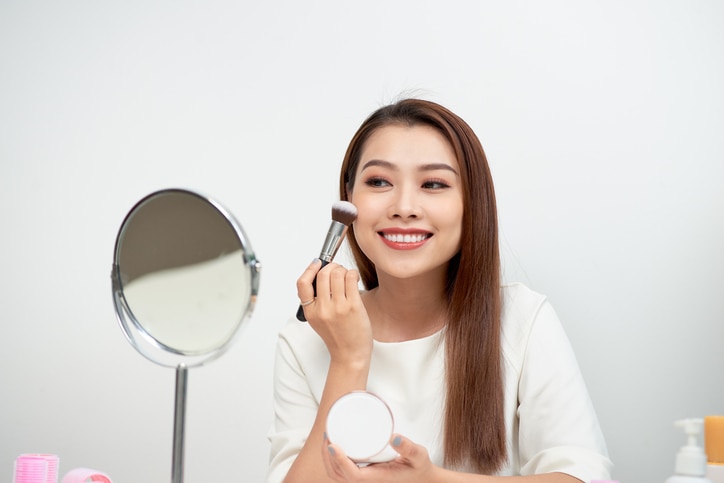 Beauty woman applying makeup. Beautiful girl looking in the mirror and applying cosmetic with a big brush. Girl gets blush on the cheekbones. Powder, rouge