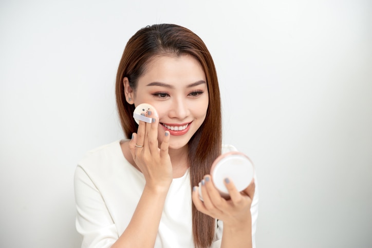 Face Make-Up. Closeup Of Sexy Female Applying Dry Powder Foundation Looking In Mirror. Portrait Of Young Woman Putting Makeup Powder With Cosmetic Cushion On Her Facial Skin Indoors. High Resolution