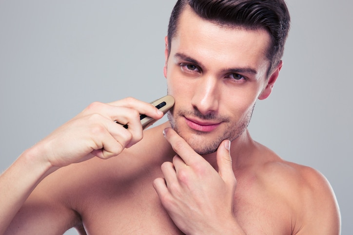 Handsome young man shaving with electric razor over gray background